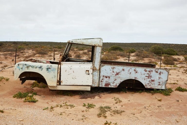 An old, rusty truck left to decay in the desert, surrounded by sand and sparse vegetation.