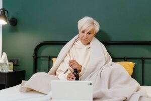 Senior woman wrapped in blanket using tablet for online medical consultation, indoors.
