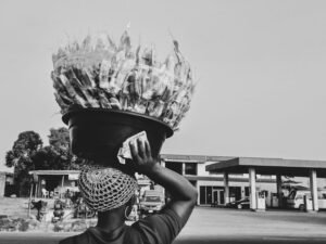 A street vendor balances a large basket of goods on their head in an urban setting.