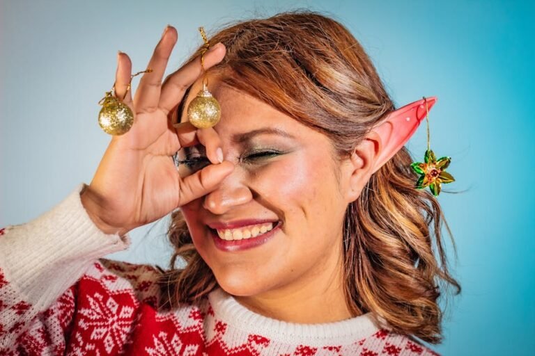 A smiling woman with elf ears playfully holding Christmas ornaments, wearing a holiday sweater.