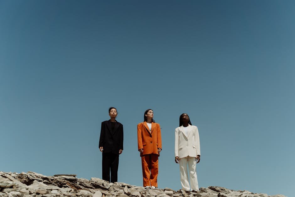 Three people in colored suits stand on rocky terrain against a clear blue sky, showcasing modern fashion.